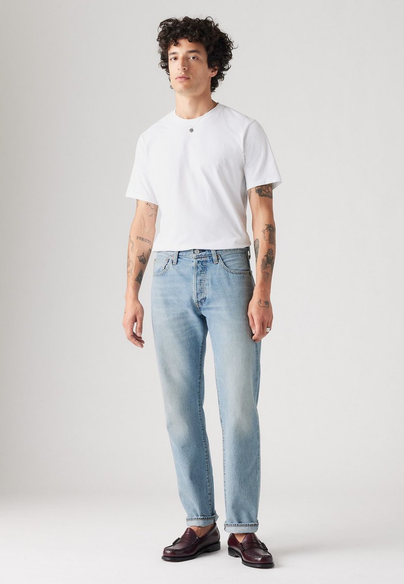Camiseta blanca de manga corta, jeans de mezclilla azul claro y mocasines burdeos oscuros. El modelo está de pie con los brazos relajados, mostrando tatuajes en los brazos.