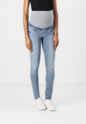 Personne vêtue d'un jean skinny de maternité bleu clair avec une ceinture élastique grise, un haut noir et des baskets blanches, debout devant un fond blanc.