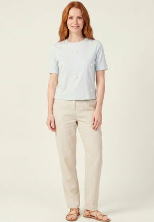Femme aux longs cheveux roux portant un haut à manches courtes bleu clair avec de petits accents blancs, un pantalon beige et des sandales marron clair, debout devant un fond uni.