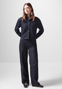 Giubbotto di denim scuro con bottoni e tasca, abbinato a pantaloni a gamba larga in denim scuro. Ballerine nere con borchie completano il look.