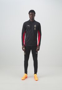 Svart atletisk träningsdräkt med röda accenter och guldlogotyp. Tätt sittande design, inkluderar zip-up jacka och smalnande byxor, kombinerad med klara gula fotbollsskor.