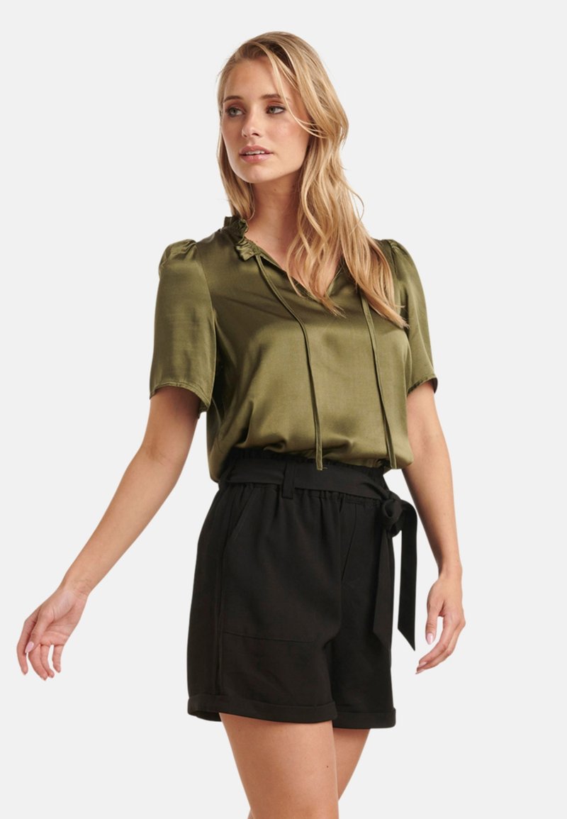 Olijfgroene satijnen blouse met korte puffmouwen en een trekkoordhals, gecombineerd met zwarte shorts met een riem en omgeslagen zomen.