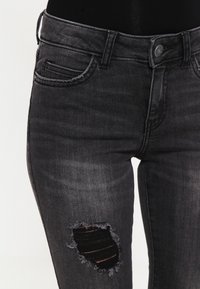 Mustat farkut, joissa on slim fit -leikkaus, revennyt polvi ja klassinen viisi-taskumuotoilu. Kangas on hieman kiiltävä.