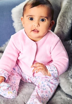 Maglione a maniche lunghe rosa chiaro in fleece con bottoni a pressione, abbinato a pantaloni rosa con motivo floreale. Bambino seduto su una superficie grigia morbida.