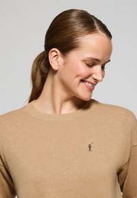 Beige gestrickter Pullover mit Rundhalsausschnitt und geripptem Muster. Mit einem kleinen gestickten Logo auf der Brust. Weicher Stoff mit lockerer Passform.