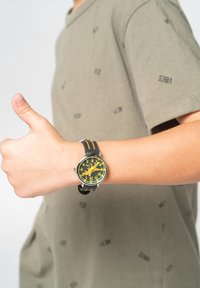 Montre ronde avec un cadran noir et jaune, grands indicateurs d'heure, et un bracelet noir avec des accents orange. Bras d'une personne levé avec un pouce en l'air.