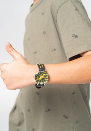 Orologio tondo con un quadrante nero e giallo, indicatori dell'ora ben visibili, e un cinturino nero con dettagli arancioni. Braccio di una persona alzato con il pollice in su.