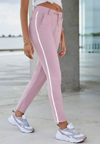 Helle rosa Sporthosen mit einem Kordelzugbund, Seitentaschen und kontrastierenden weißen Seitenstreifen. Kombiniert mit grau-weißen Sneakers.