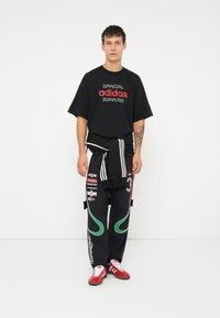 Melns T-krekls ar sarkano "adidas" logo, brīvs piegriezums. Tumši bikses ar zaļiem un baltajiem akcentiem, teksta grafikām. Sarkanas kedas ar baltām svītrām.