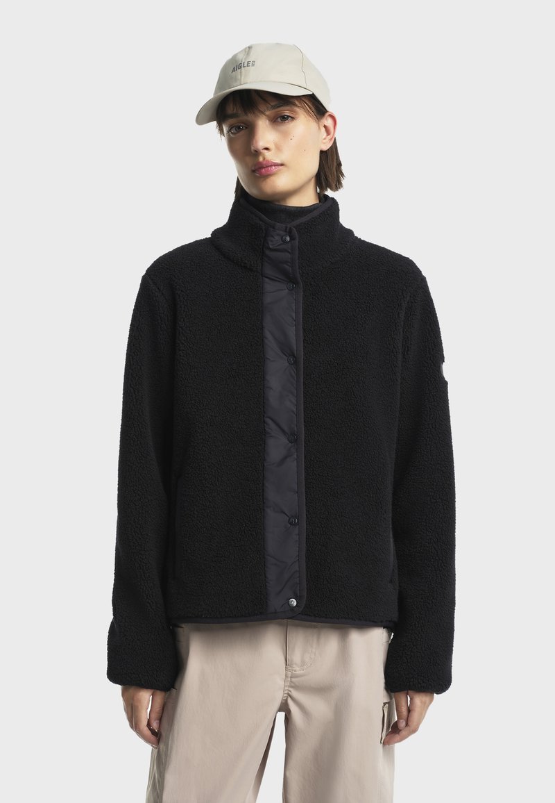 Veste en polaire noire avec un col montant et une fermeture à bouton-pression, présentant un panneau lisse contrastant. Portée avec un pantalon clair et une casquette beige.