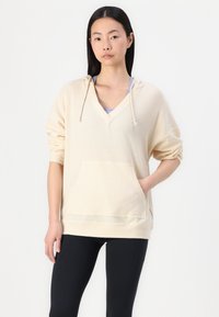 Beige Kapuzenpullover mit V-Ausschnitt, strukturiertem Stoff, Fronttasche und umgekrempelten Ärmeln. Kombiniert mit schwarzen Leggings.
