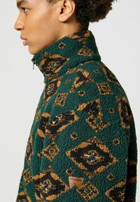 Sudadera de forro polar verde con patrón geométrico en marrón y negro, cuello alto, cierre completo y pequeño parche del logo de Wrangler en el lado izquierdo.