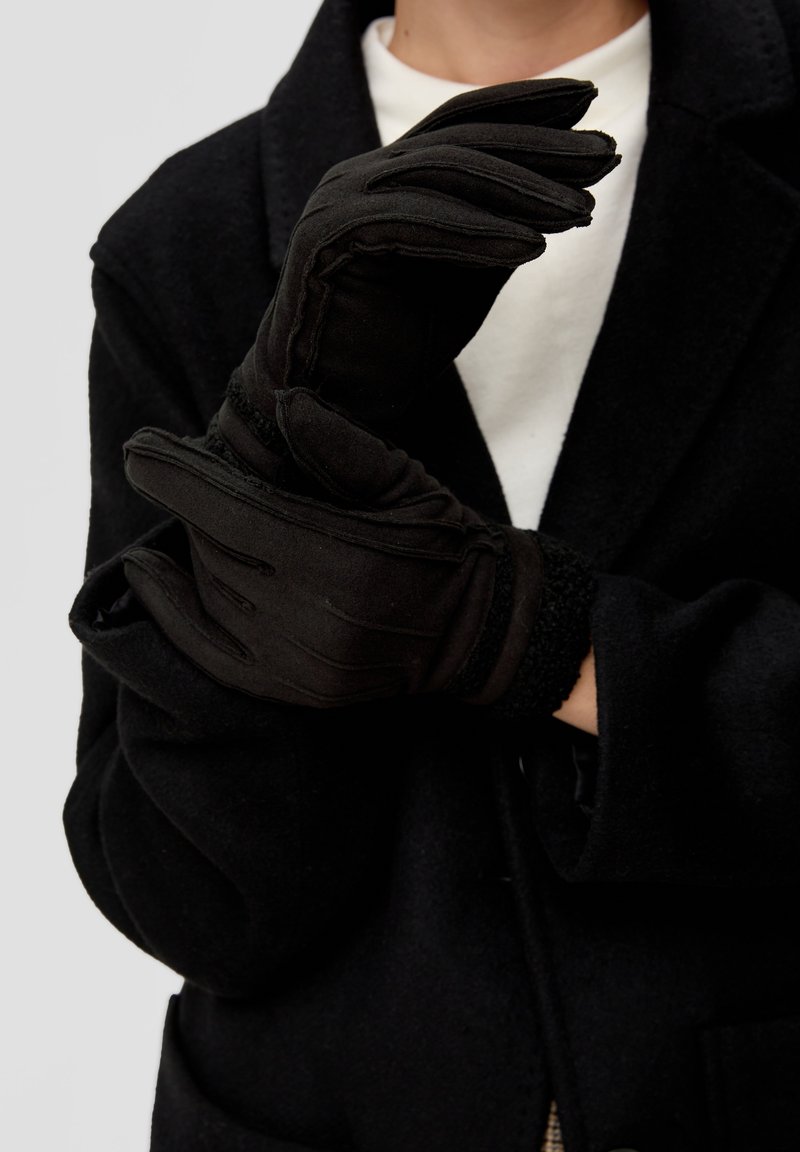 Personne portant des gants noirs et un manteau noir, ajustant un gant avec l'autre main sur un fond uni.