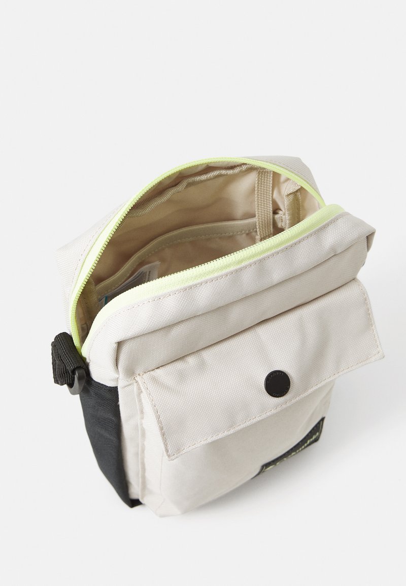 Pequeño bolso cruzado beige y negro con un compartimento principal con cremallera abierta y un bolsillo frontal con botón a presión sobre un fondo blanco.