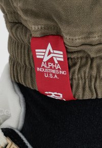 Tejido color caqui con textura acanalada, que presenta una gran etiqueta roja con texto blanco que dice "ALPHA INDUSTRIES INC. U.S.A." y acentos de logotipo.