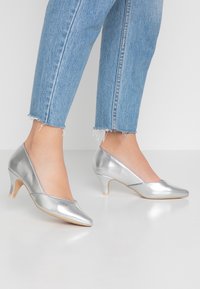 Talons argentés avec un bout pointu et un design en V. Matériau métallique lisse, talon slim de 5 cm et esthétique minimaliste.