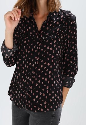 Blouse noire à taches abstraites roses, détails volantés sur les épaules et la poitrine, portée avec les manches retroussées et un jean gris foncé.