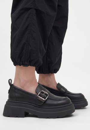 Zwarte plateau loafers met zilveren gespdetail, gedragen met zwarte opgerolde broeken, waarbij de enkels zichtbaar zijn tegen een effen lichte achtergrond.