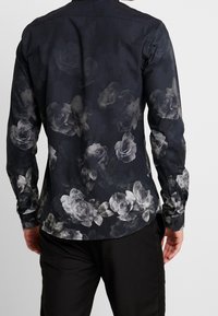 Maglietta a maniche lunghe realizzata in un tessuto scuro con un grande motivo floreale grigio, orlo arrotondato e polsini con bottoni. Aspetto texture.