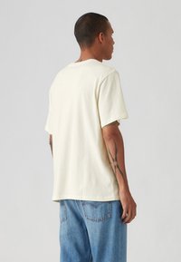 Crème-kleurige t-shirt met korte mouwen, gemaakt van zacht materiaal, met een ontspannen pasvorm en een ronde zoom. Zichtbare tatoeages op de arm.