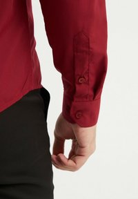 Chemise rouge à manches longues avec poignets à boutons, présentant une texture lisse et une coupe ajustée. Comprend deux boutons au poignet et un détail de manches pointues.