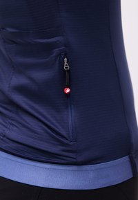 Krupni plan tamnoplave teksturirane odjevne tkanine s džepom na patentni zatvarač koji ima crnu traku za povlačenje i crveni ukras u obliku perlice, nošene preko tamnih hlača.