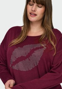 Burgunderfarget langermet topp med et stort sølv-leppeprint laget av små prikker. Myk tekstur med en avslappet passform og bred hals.