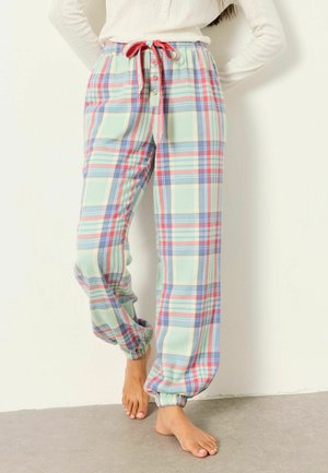 Vrouw draagt pastelgeruite pyjamabroek met elastische boorden en een voorstrik, staat blootsvoets op een grijze vloer.