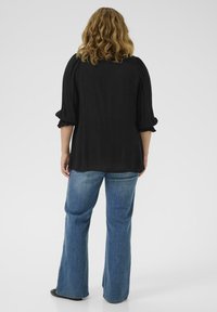 Black, long-sleeve top with ruffled cuffs, paired with light blue flared jeans; model is seen from the back against a plain background.