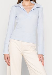 Haut à manches longues bleu clair avec un col en V et un large col, fabriqué en tissu côtelé. Porté avec un pantalon à jambe large de couleur crème.