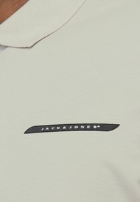 Lys grå polo skjorte med struktureret strik, der har et sort broderet "JACK & JONES" logo på venstre side. Klassisk krave design.