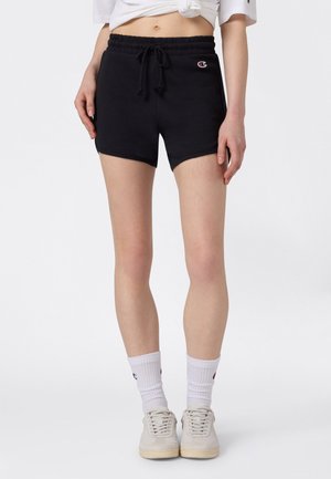 Kobieta w czarnych krótkich spodenkach z sznurkiem, białych skarpetach do połowy łydki, białych sportowych butach i skróconym białym topie na jednolitym tle.