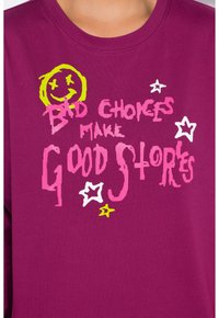 Sweatshirt magenta arboré d'un texte graphique : "LES MAUVAISES CHOIX FONT DE BONNES HISTOIRES," accompagné d'un visage souriant et d'étoiles en jaune, rose et blanc.