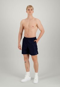 Costume da bagno blu navy con elastico in vita e tasca laterale. Indossato con scarpe da ginnastica bianche e calze bianche. Tatuaggi visibili sulla gamba sinistra.