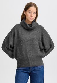 Maglione grigio oversize con collo alto, realizzato in un tessuto a maglia testurizzata, con spalle scese e un ampio colletto ripiegato. Abbinato a jeans blu.