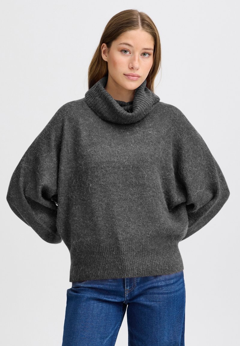 Maglione grigio oversize con collo alto, realizzato in un tessuto a maglia testurizzata, con spalle scese e un ampio colletto ripiegato. Abbinato a jeans blu.