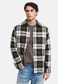 Koszula flanelowa w kratę w kolorach zielonym, białym i pomarańczowym. Posiada dwie kieszenie na piersiach oraz kołnierz z guzikami, nałożona na brązowy sweter.