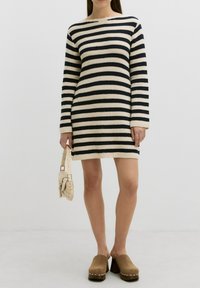 Gestreift, gestricktes Kleid in Creme und Marine mit langen Ärmeln, einem U-Boot-Ausschnitt und einem hohen Saum. Abgerundet mit einer beigen Schultertasche und Clogs.