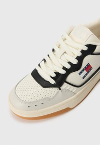 Cream and black leather sneaker with perforated toe, textured accents, and a gum rubber sole. Features Tommy Jeans logo and flat laces.