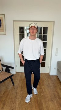 Mladý muž oblečený v bielom tričku, námorníckych modrých nohaviciach, bielych teniskách a šiltovke jachtárskeho klubu stojí vo vnútri s rukami v vreckách.