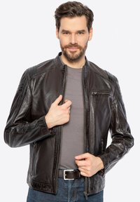 Giubbotto in pelle marrone con chiusura a zip, colletto rialzato, accenti sulle spalle e tasche laterali, indossato sopra una maglietta grigia e jeans blu.