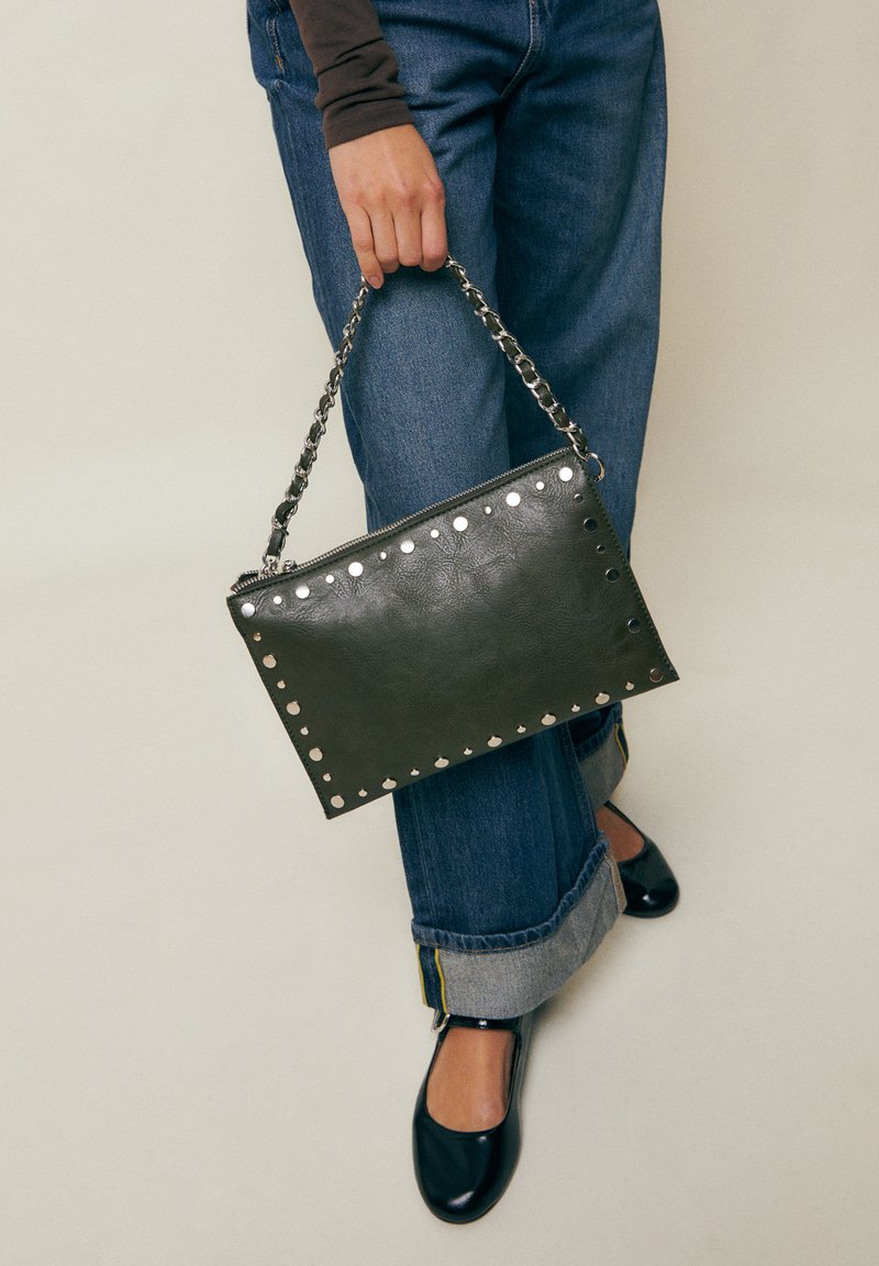 Isik, kes hoiab musta nahast käekotti, millel on hõbedased naelad ja kettist rihm, kannab pööratud äärtega siniseid teksapükse, musti jalatseid ja pika varrukaga pluusi.