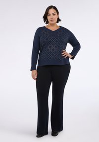 Tmavě modrý top s dlouhým rukávem, zdobený diamantovým vzorem a výstřihem do V. V kombinaci s černými rozšířenými kalhotami a černými botami.