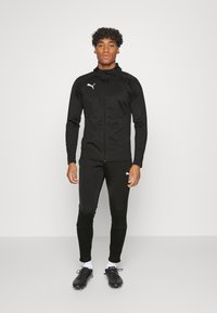 Musta urheilu takki vetoketjulla, jossa on korkea kaulus ja valkoinen logo. Yhdistetty mustiin sovittimiin ja mustiin juoksukenkään.