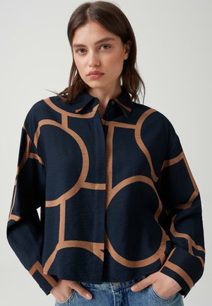 Femme portant une chemise bleu marine avec de grands motifs géométriques circulaires et linéaires beige, debout les mains dans les poches devant un fond uni.