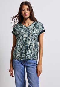 Grünfarbener Kurzarm-Top mit V-Ausschnitt und schwarzem Schlangenprint-Muster. Weicher Stoff mit umgeschlagenen Ärmeln und lockerer Passform. Kombiniert mit blauen Jeans.