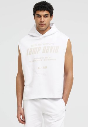 Man wearing sleeveless white hoodie with hood and light text, paired with white pants, standing against plain light background.