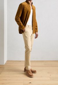 Camisa marrón de pana con botones, camiseta interior blanca, pantalones beige y mocasines marrones de ante. Atuendo casual con tela texturizada.