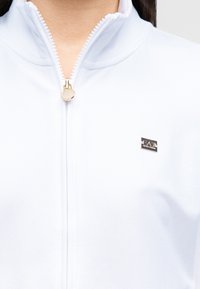 Λευκό μπουφάν με φερμουάρ και χρυσό γαντζάκι φερμουάρ, με μικρή ορθογώνια κονκάρδα λογότυπου EA7 στο αριστερό στήθος, φορεμένο από ένα άτομο.