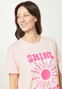 Lyserød t-shirt i blød stof med et markant, pink solgrafik og teksten "SHINE". Korte ærmer med opsmøg.